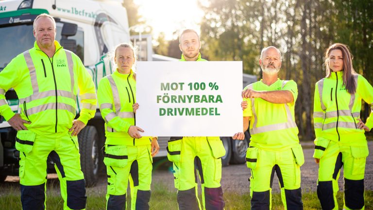 Fem personer i varierande åldrar, tre män och två kvinnor, står framför en grön och vit lastbil - LBC Frakt. De har gula varselkläder på sig och de håller i en skylt med texten: Mot 100% förnybara drivmedel