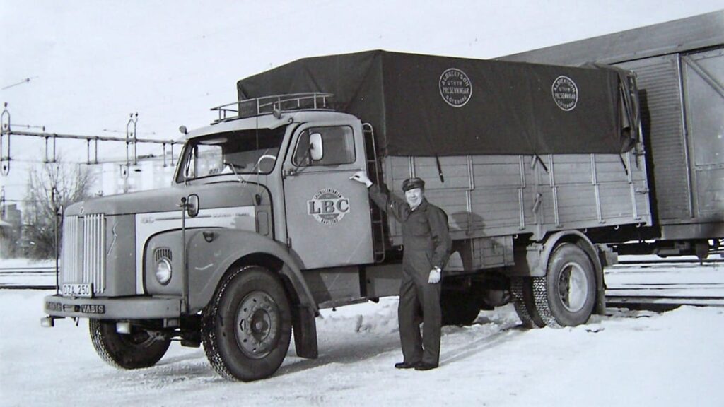 En svartvit bild på en gammal lastbil intill en tågvagn och en man som står vid förarsidan och tittar mot kameran. Det är vinter och snö på marken.