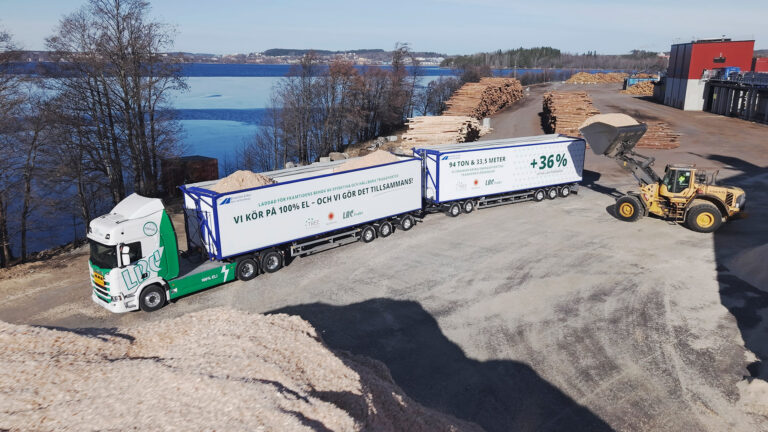 Bil på en lång lastbil från LBC Frakt. Lastbilen står parkerad på en grusväg vid en sjö och blir lastad med flis. På lastbilen står: Vi kör på 100% el-och vi gör det tillsammans (Stora Enso, TREE, LBC Frakt). 94 ton, 33,5 meter, +36%