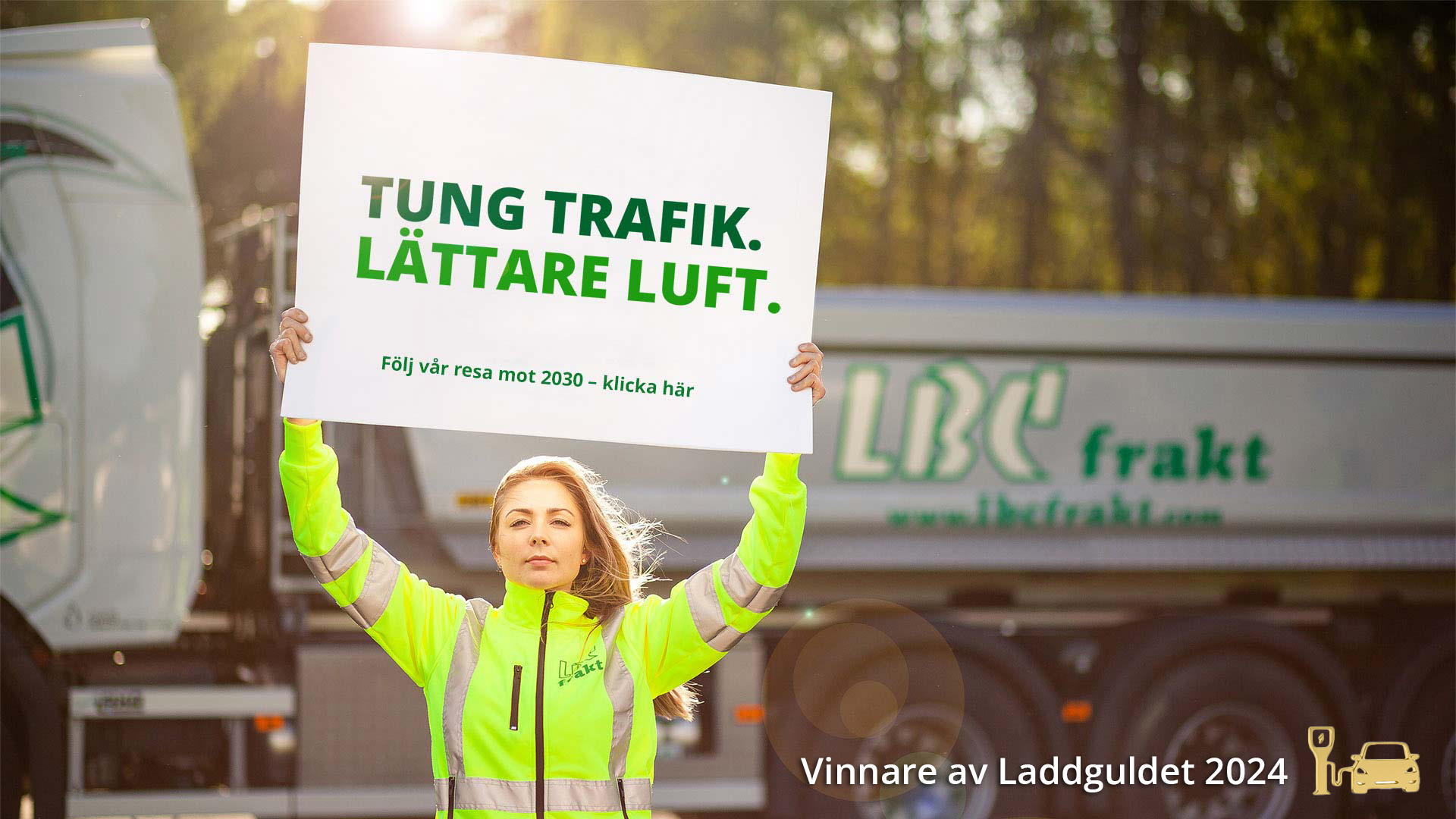 Bild på en kvinnlig chaufför som håller en skylt med texten: TUNG TRAFIK LÄTTARE LUFT Följ vår resa mot 2030- klicka här. Och texten: Vinnare av Laddguldet 2024 + loggan för Laddguldet (en bil som laddar vid en stolpe. loggan är i guld).