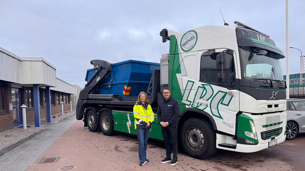 Bild på en eldriven kroklastbil från LBC Frakt. På lastbilen står en blå container. Framför lastbilen står vd för Rydahls reservdelar och affärsområdeschef för Bygg, anläggning & miljö. De ser glada ut.