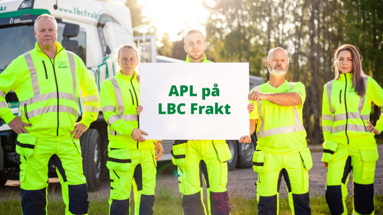 Fem chaufförer klädda i gula varselkläder håller i en skylt med texten "APL på LBC Frakt". Chaufförerna är i olika åldrar, tre män och två kvinnor. I Bakgrunden står en av LBC Frakts lastbilar.